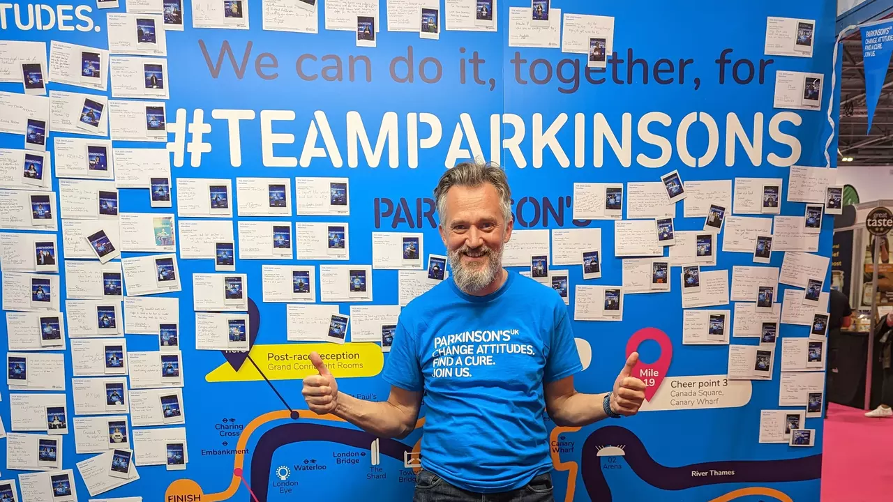 A man standing in a cyan blue t-shirt standing in front of a cyan blue board that reads 'Team Parkinson's'