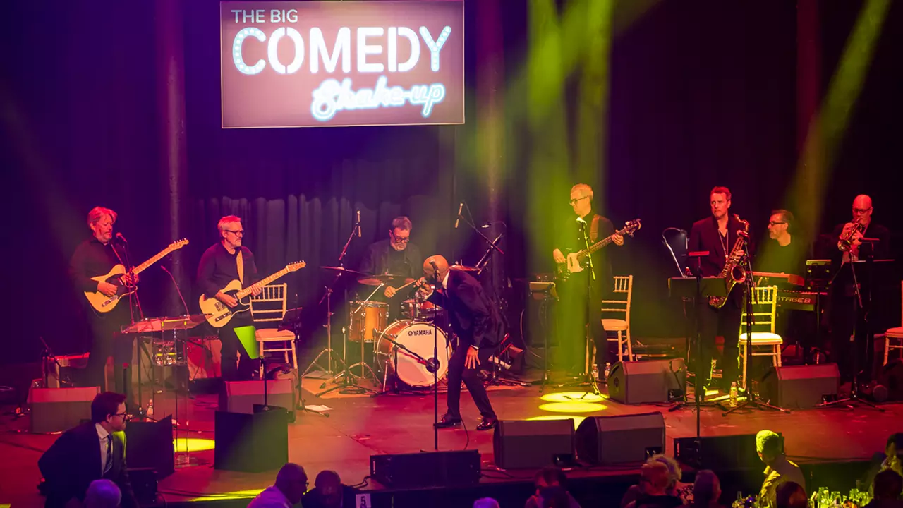 A band on stage with the big comedy shake up logo behind them