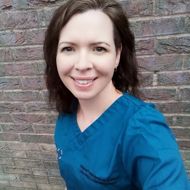 Nic is wearing a blue scrub top and smiling to camera. She has shoulder-length brown hair and is stood in front of a brick wall.