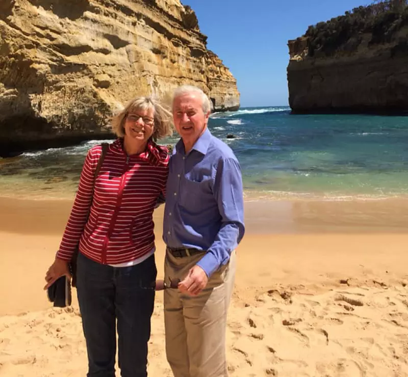 Jim and Jane are standing on a beach. Jane is wearing a red jumper and blue jeans. Jim has a blue shirt on, with beige chinos. They are both smiling. 