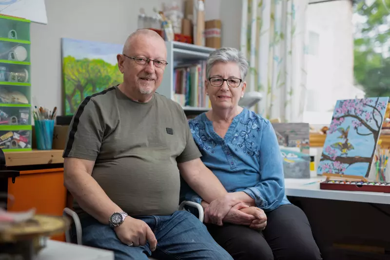 John and Susan are sittng down, holding hands. They are surrounded by their artwork.