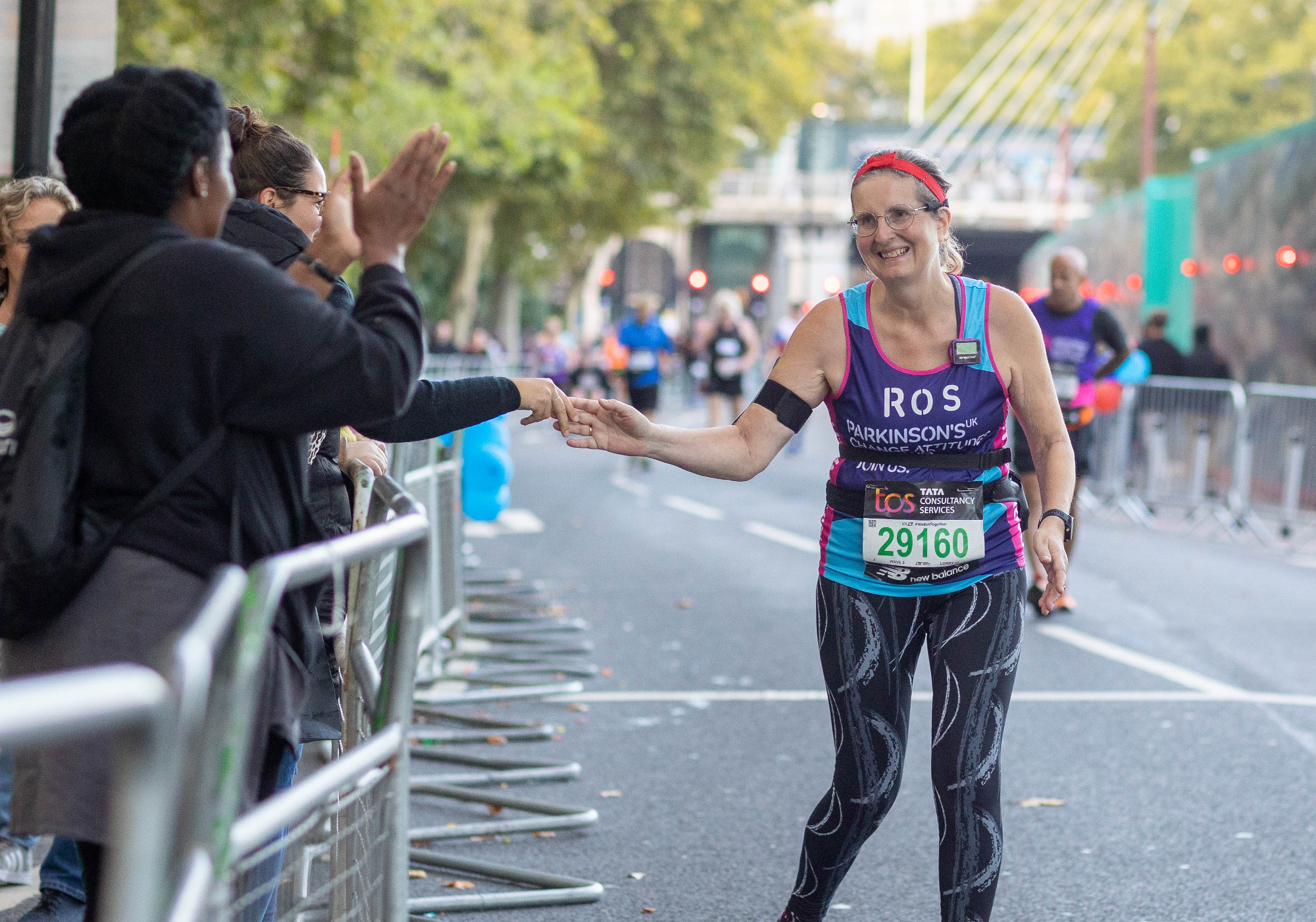 An older woman running in a Parkinson's UK running top that has the name 'Ros' on it. She is smiling and brushing fingers with someone who is watching from the sidelines.