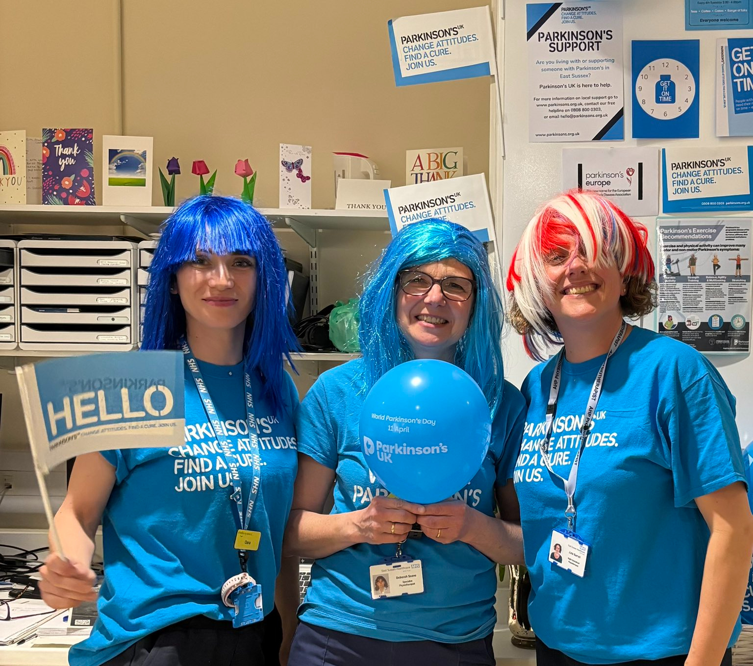 Three women wearing fancy dress wigs and Parkinson's UK tshirts hold balloons and flags.