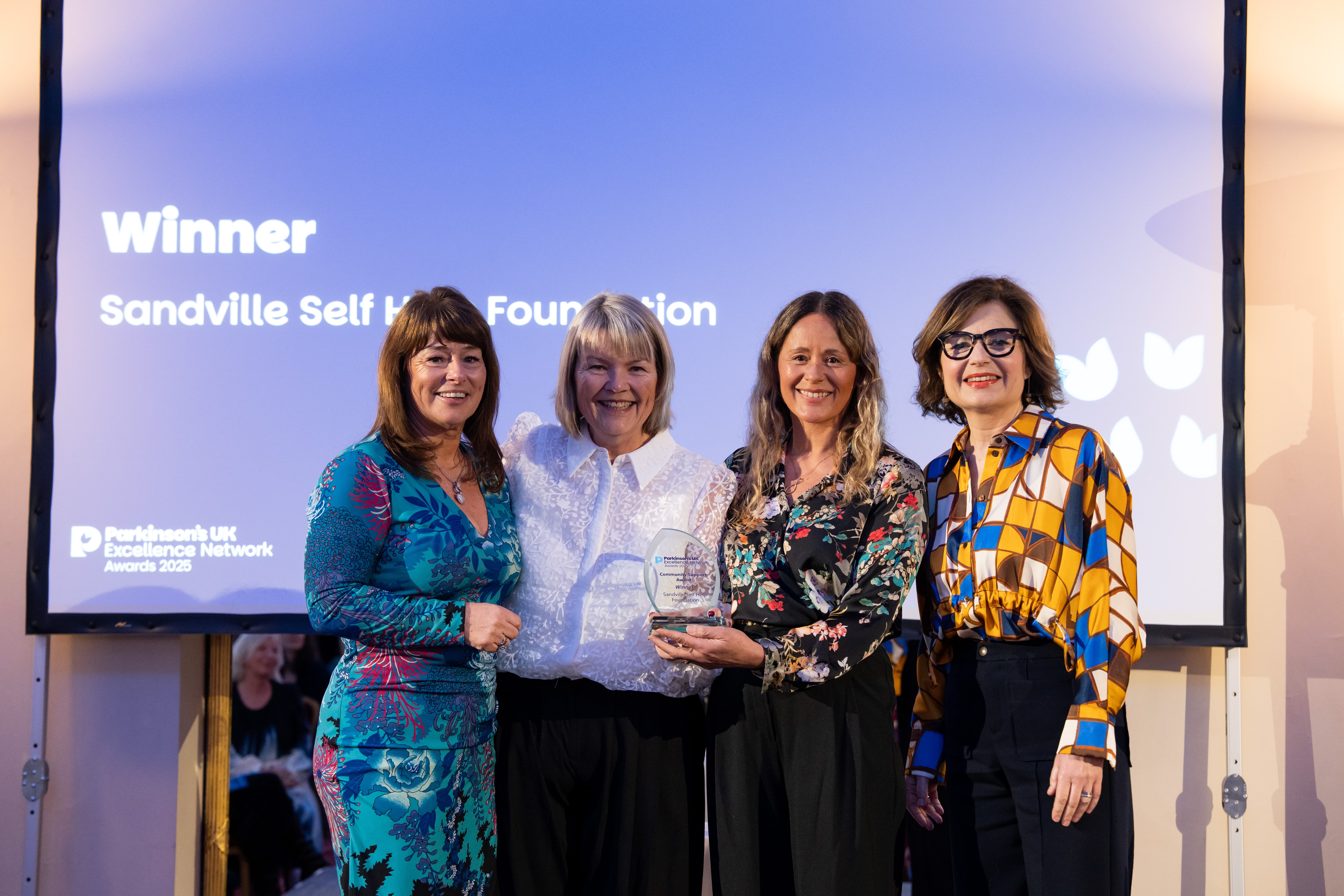 Four women stand in front of a screen showing the word "Winner". They are smiling.