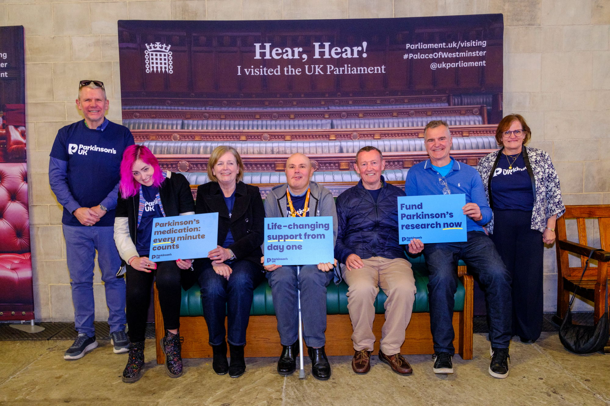A group of men and women sitting on a bench in Parliament. They are holding signs that say 'Parkinson's medication: every minute counts', 'Life-changing support from day one' and 'Fund Parkinson's research now'.
