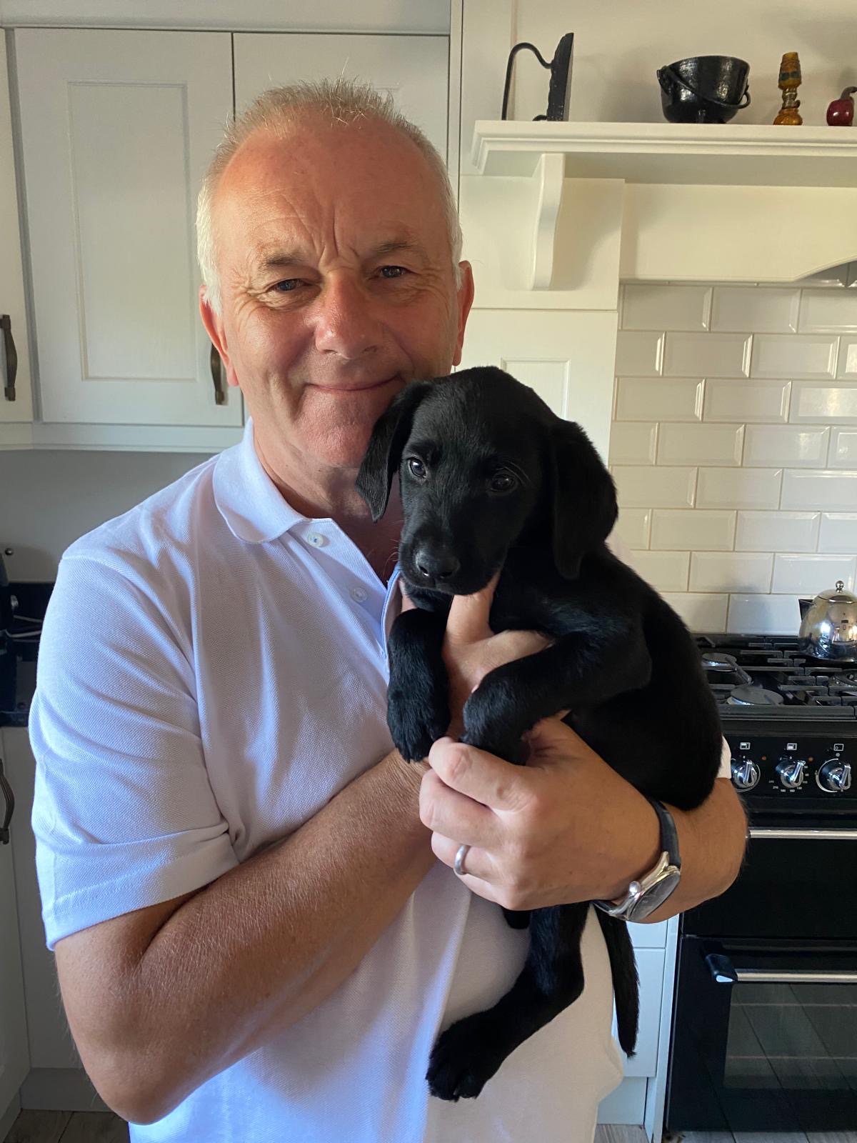 A white man in a white shirt is holding a small, black Labrador puppy and smiling into the camera. 