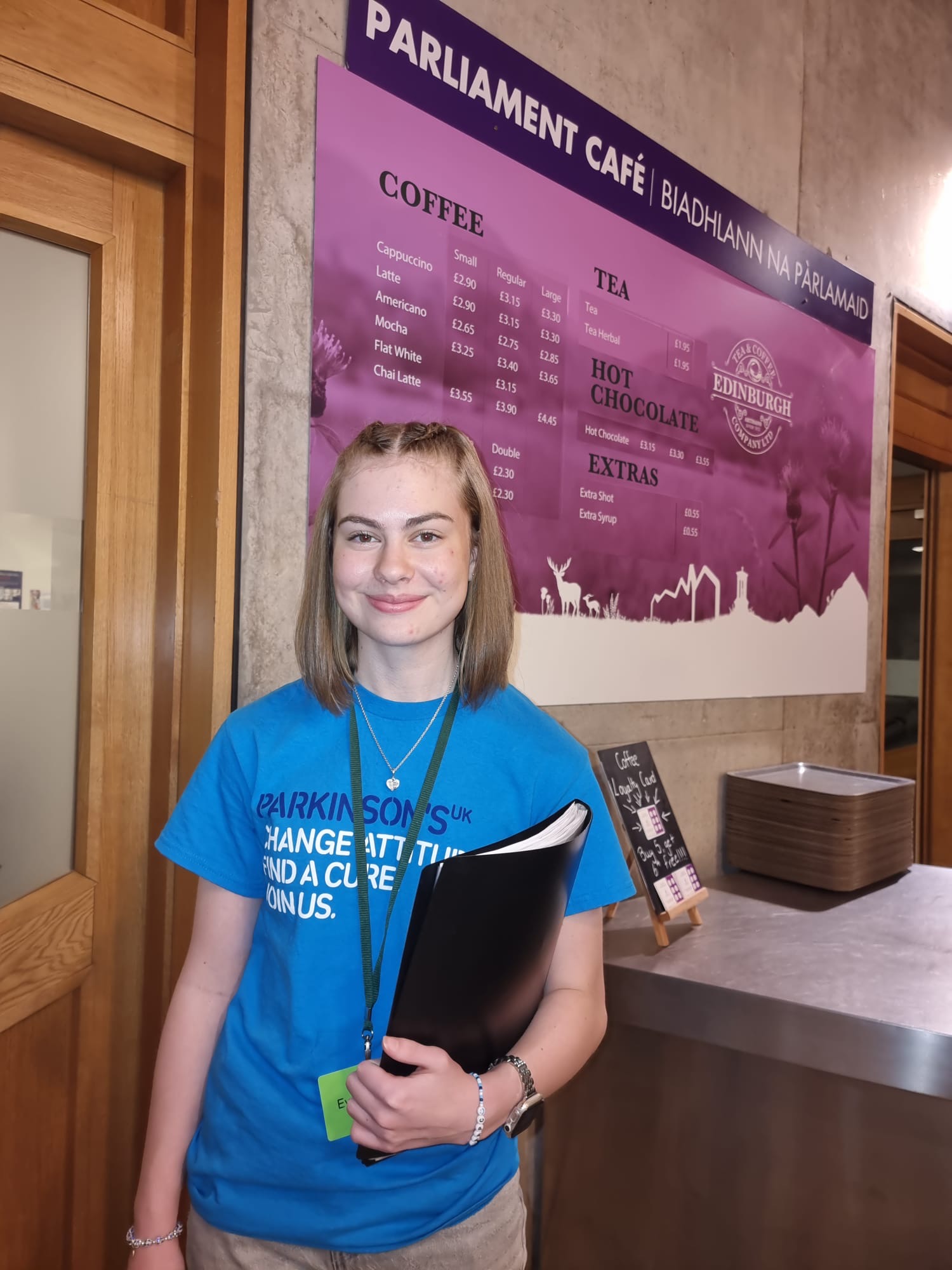 A young white lady smiles for the camera wearing a blue Parkinson's UK t-shirt and holding a black folder. 