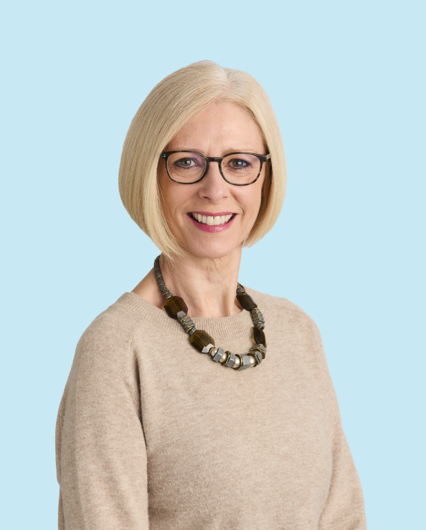 A white woman with a blonde bob, glasses and wearing a beige jumper is looking into the camera and smiling for a professional headshot.