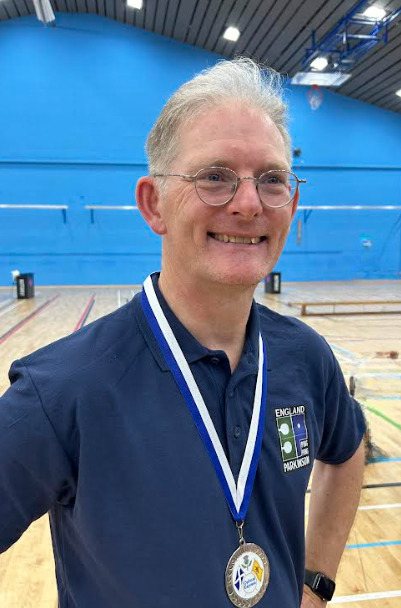 A man wearing glasses a blue polo shirt and a medal stands proudly and smiles.