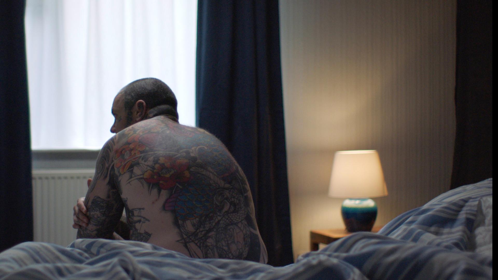 Shaun, a young and heavily tattooed man with Parkinson's, site on his bed, facing away from the camera