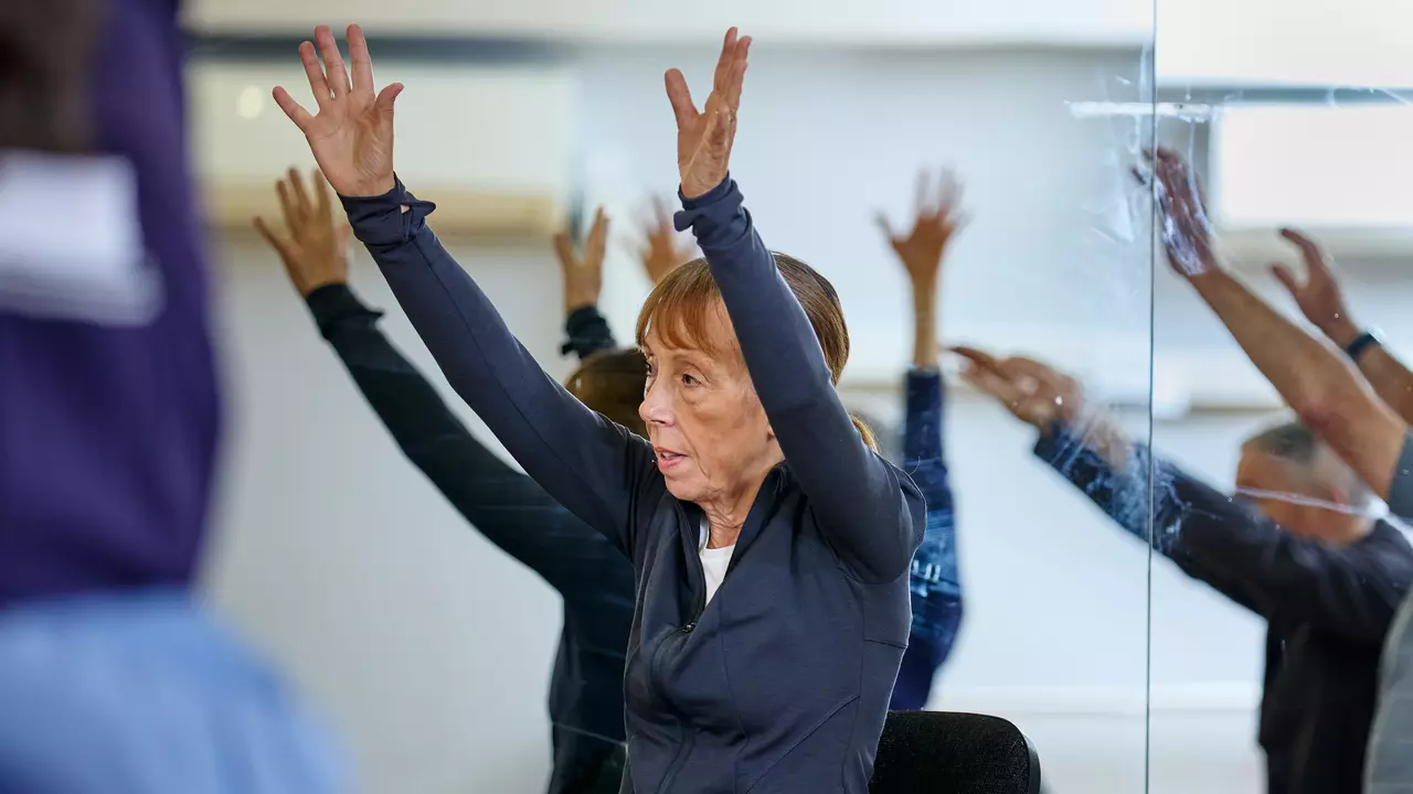 An image of a woman in a seated exercise class. The woman is raising both her arms in the air for a stretching routine.
