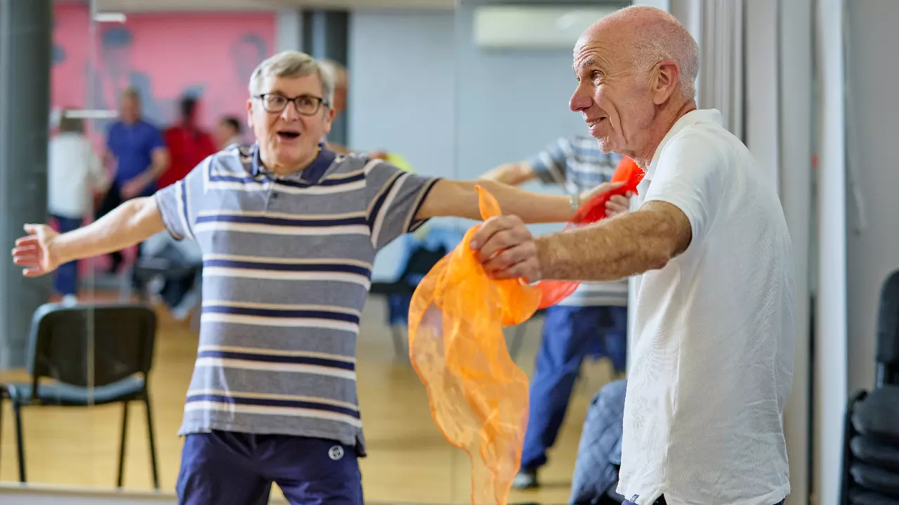 An image of two men with Parkinson's taking part in a Parkinson's exercise class. One is stretching his arms and holding an orange ribbon while the other is stretching their arms and singing loudly