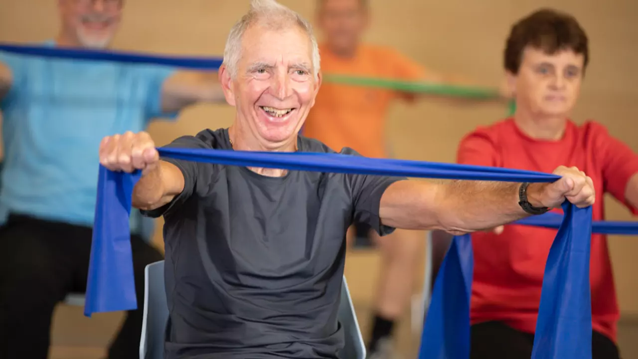An image of a man in a seated exercise class, he is stretching a resistance band with other members of the class, while in a seated position. he is wearing a dark grey top and dark grey shorts while holding a dark blue resistance band.