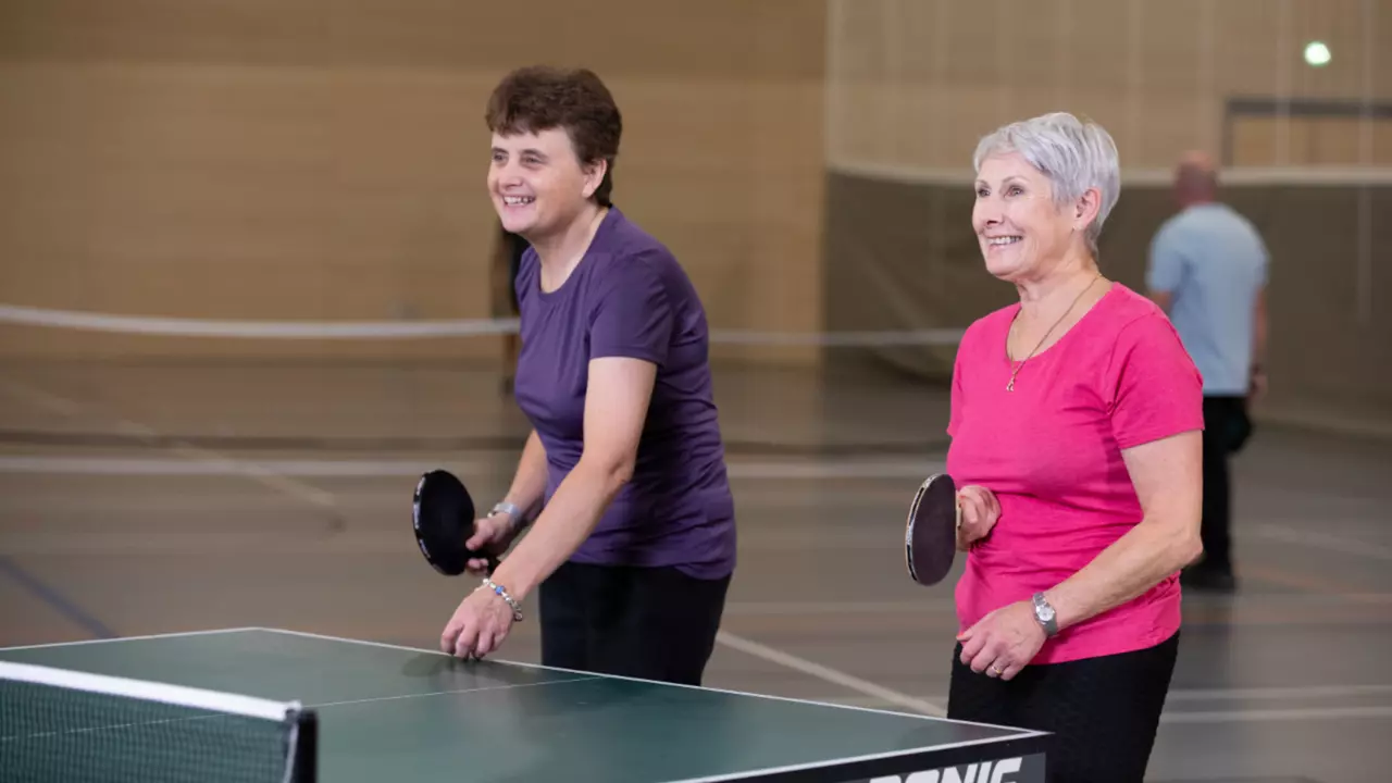 An image of two women with Parkinson's playing table tennis. One woman is wearing a purple top with black trousers and the other woman is wearing a pink top and black trousers