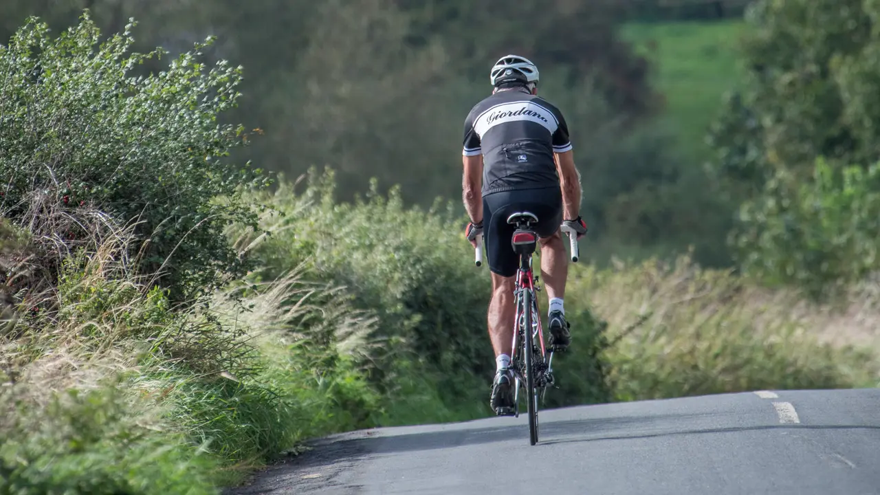 An image of a man cycling away from camera on a road bike. He is wearing a cycling helmet, protective knee and elbow guards, black shorts, a black top and special cycling shoes. 