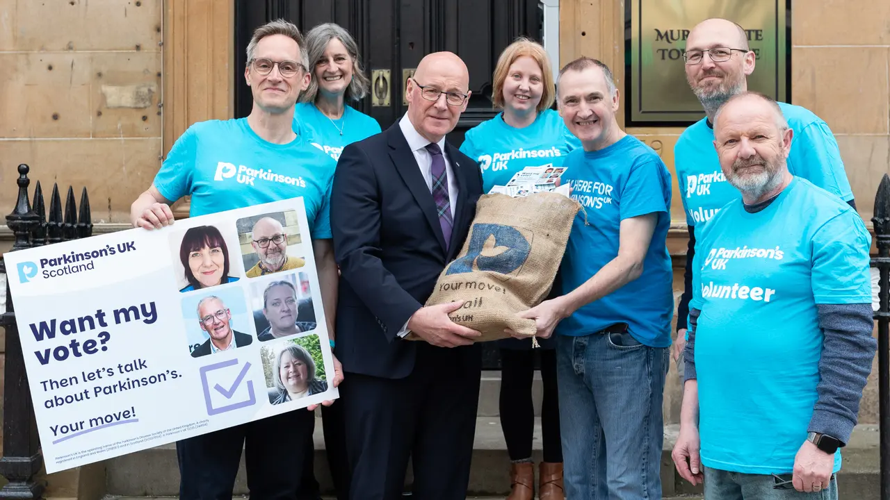 A group of people in Parkinson's UK t-shirts stand on the steps with First Minister, John Swinney. John is carrying a sack that says 'Your move! Mail' which is filled with postcards. Another member of the group is holding an oversized postcard that says 'Want my vote? Then let's talk about Parkinson's. Your move!'.