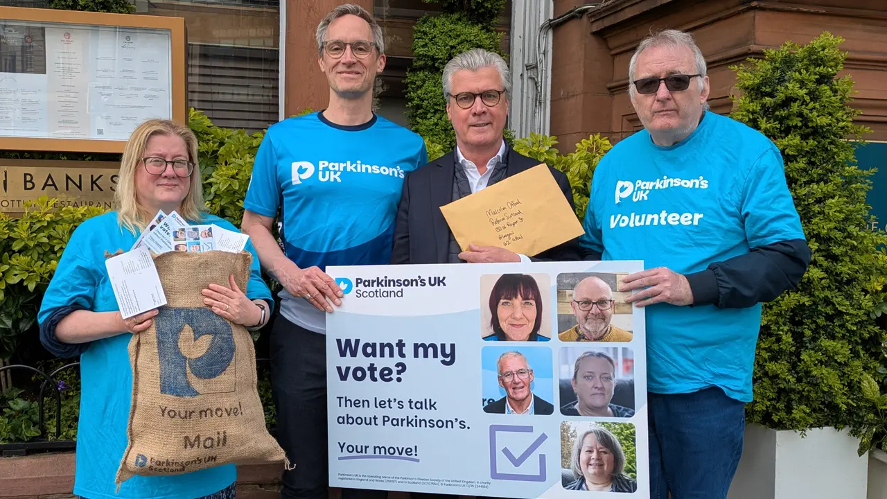 3 people in Parkinson's UK t-shirts stand with Reform UK Scotland leader, Malcolm Offord. Together, they're holding a mailbag overflowing with 'Your Move' postcards, and a oversized postcard that says 'Want my vote? Then let's talk about Parkinson's. Your move!'.