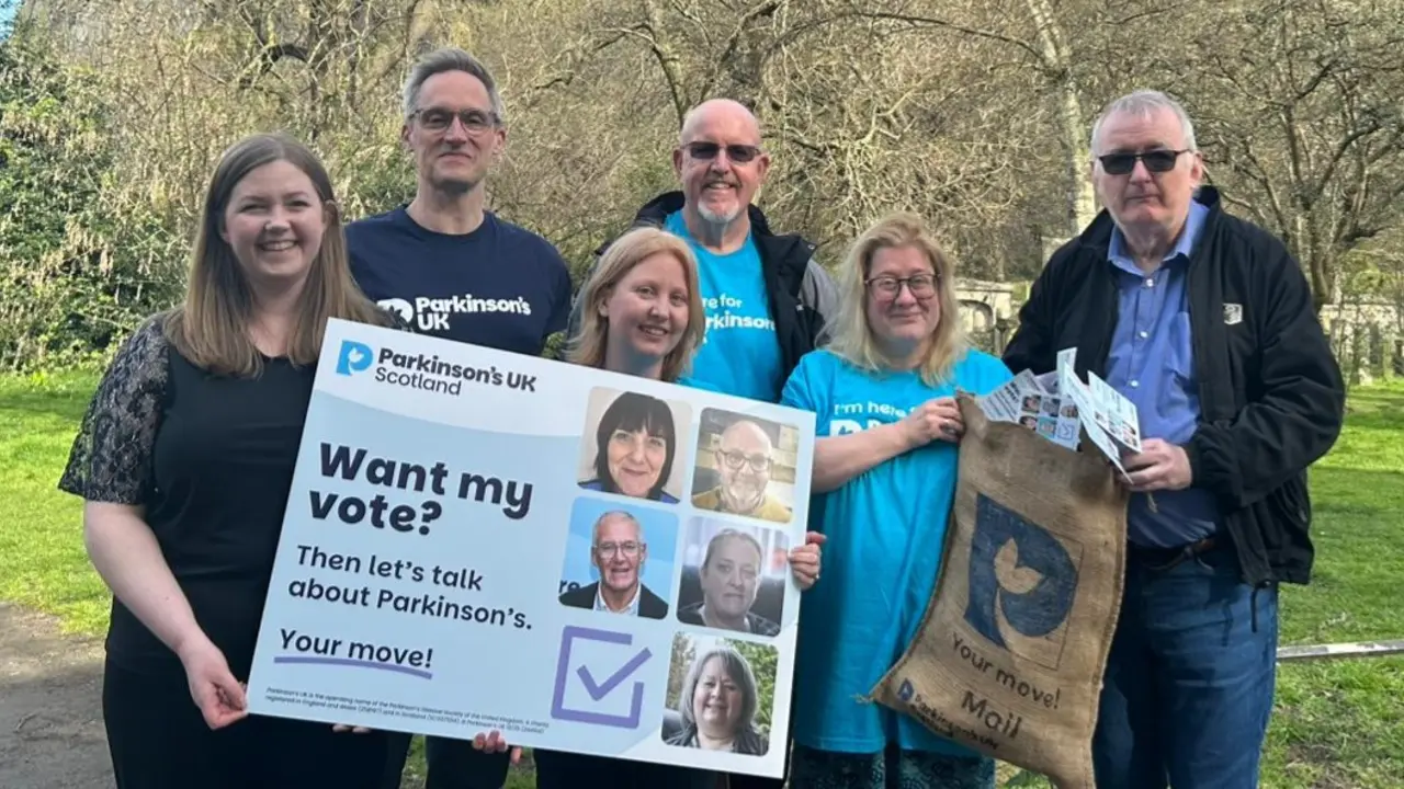 A group of people in Parkinson's UK t-shirts stand with Scottish Green co-leader, Gillian Mackay. Together they're holding a sack that says 'Your move! Mail' which is filled with postcards and an oversized postcard that says 'Want my vote? Then let's talk about Parkinson's. Your move!'.