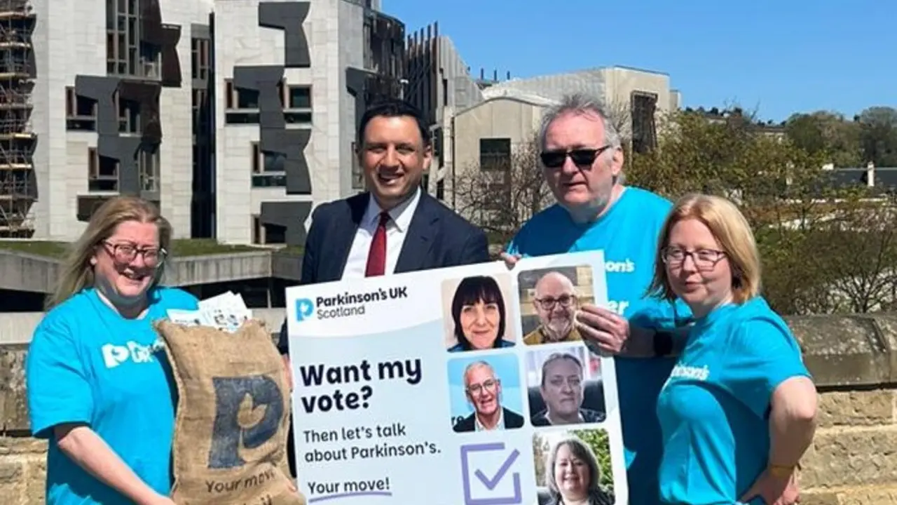 3 people in Parkinson's UK t-shirts stand with Scottish Labour leader, Anas Sarwar. Together they're holding a sack that says 'Your move! Mail' which is filled with postcards and an oversized postcard that says 'Want my vote? Then let's talk about Parkinson's. Your move!'.