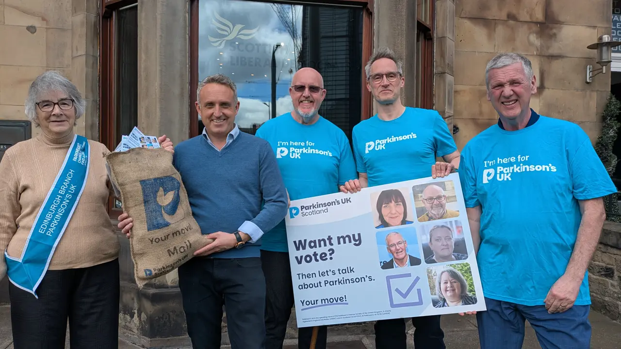 A group of people in Parkinson's UK t-shirts standing with Scottish Liberal Democrat leader, Alex Cole-Hamilton. Together they're holding a mail bag overflowing with 'Your move' postcards and an oversized postcard that says 'Want my vote? Then let's talk about Parkinson's. Your move!'.