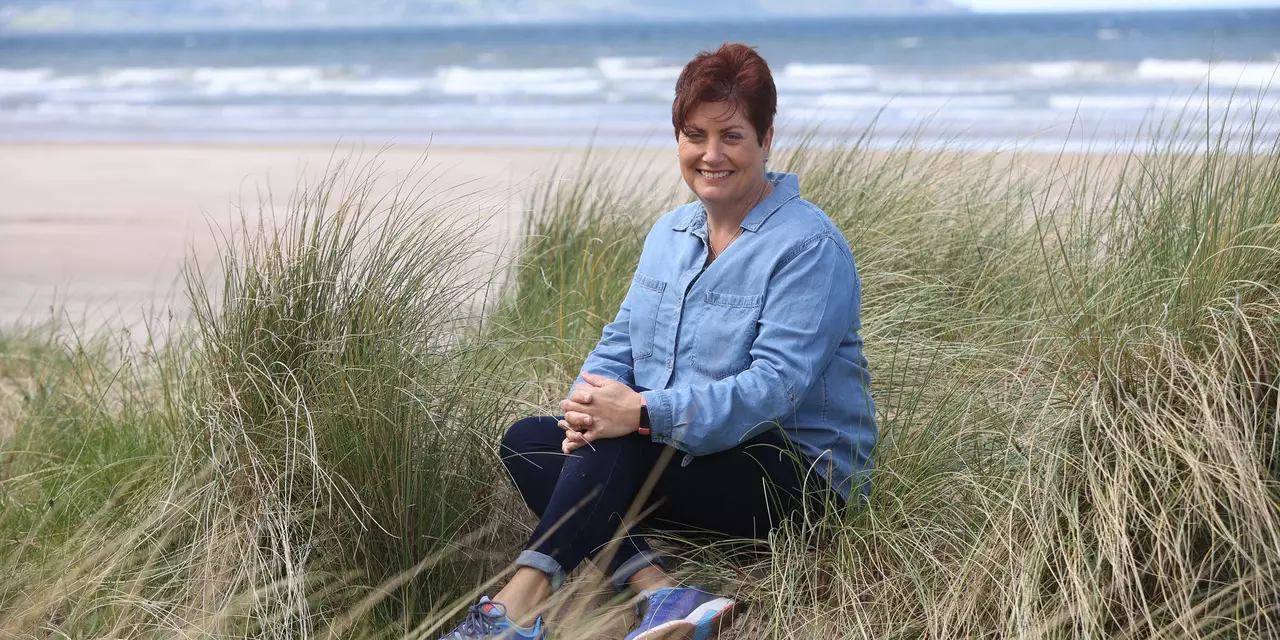 Elaine sat on the beach. She is wearing denim jeans and a denim shirt. She has short, brown hair.