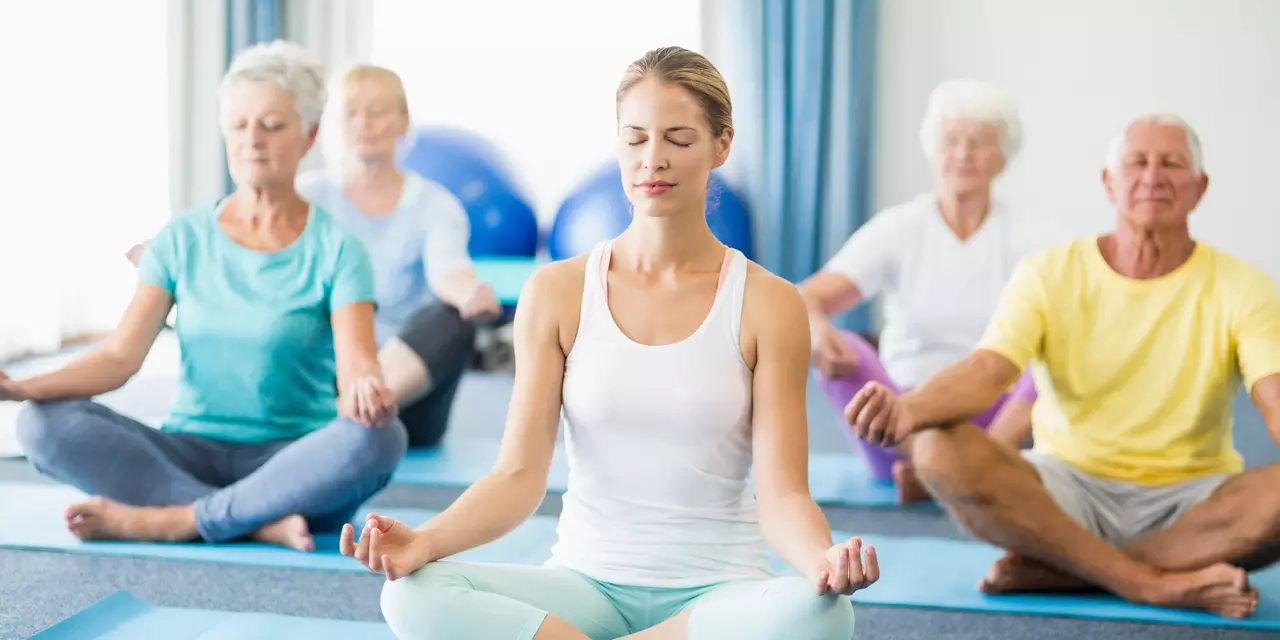 An image of an indoor yoga class, featuring a young yoga instructor and a group of people practicing yoga on mats
