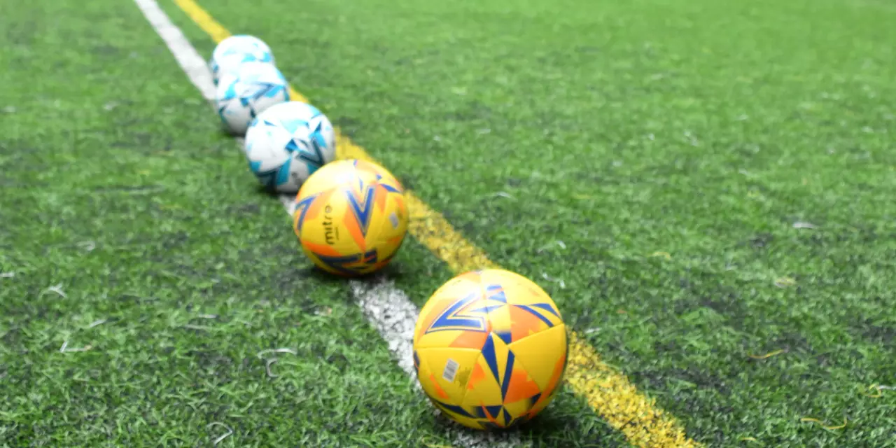 An image of an indoor football pitch with a set of six footballs. three balls are bright yellow and the other three are white. The pitch is made from green astro-turf