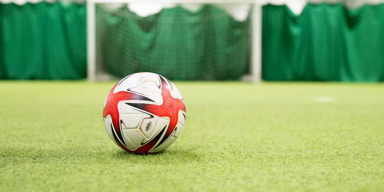 An image of a white Adidas football with a red and black cross design on a green indoor astro-turf pitch. In the background there is an empty football goal and net in white. 