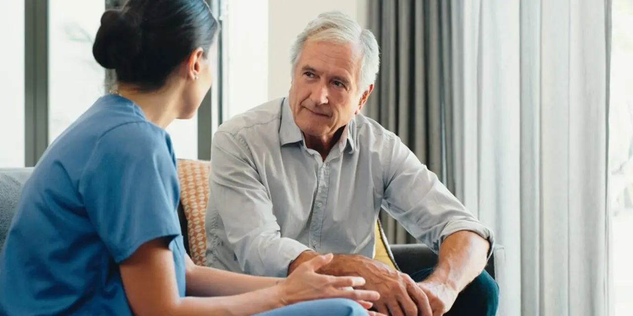 A seated older white man speaks with a nurse whose back is to the viewer.