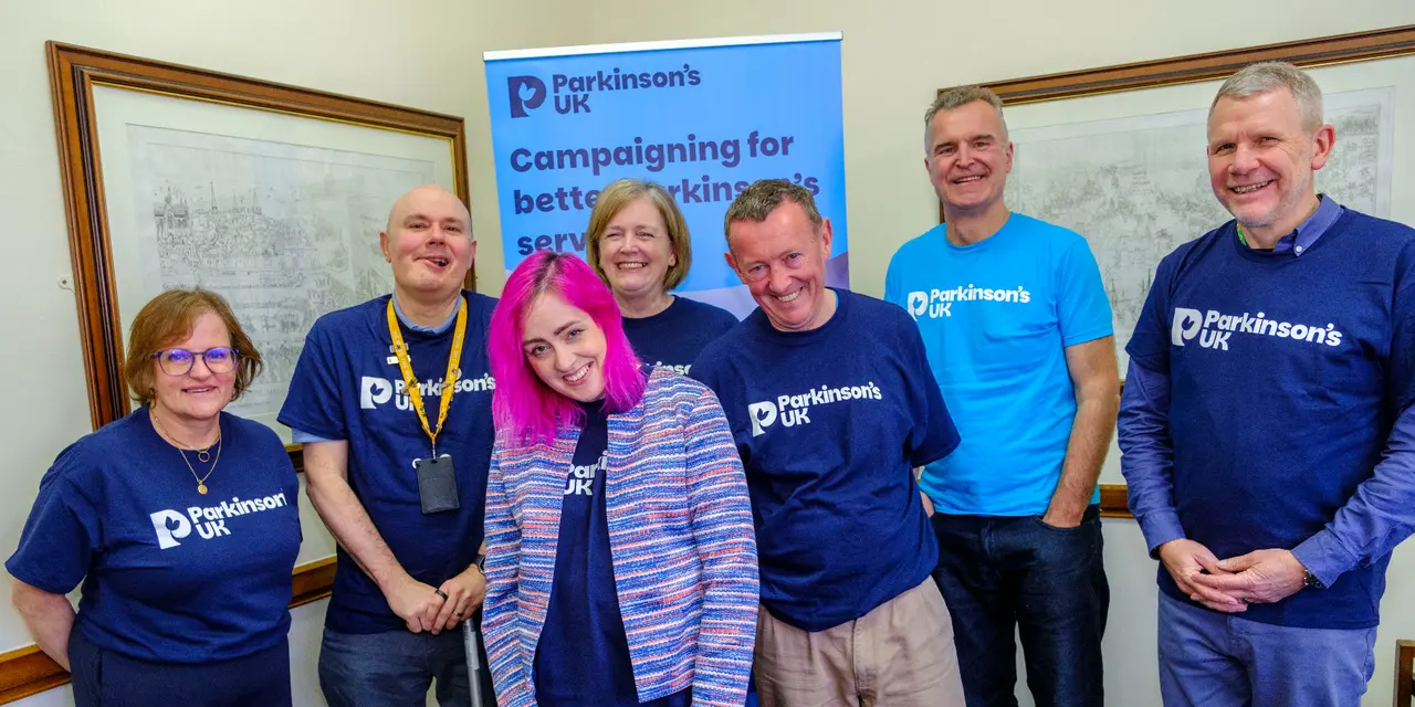 A group of smiling men and women wearing Parkinson's UK t-shirts infront of a banner that says 'Campaigning for better Parkinson's services'.