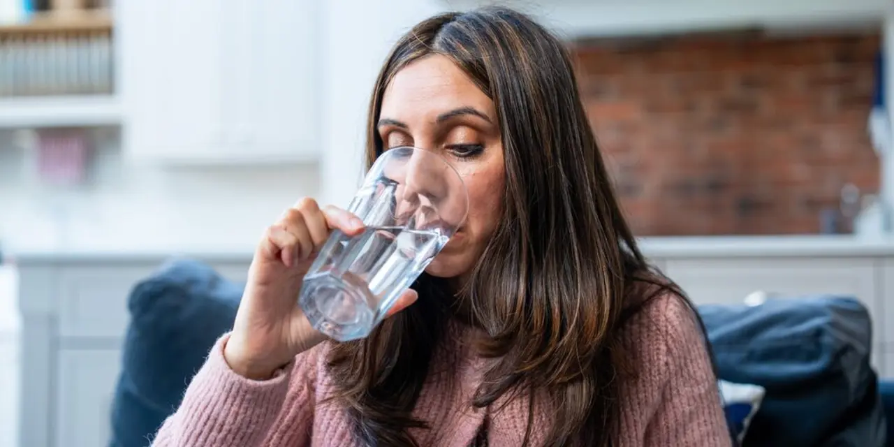 A Sourh Asian woman sits on a sofa and drinks water. 
