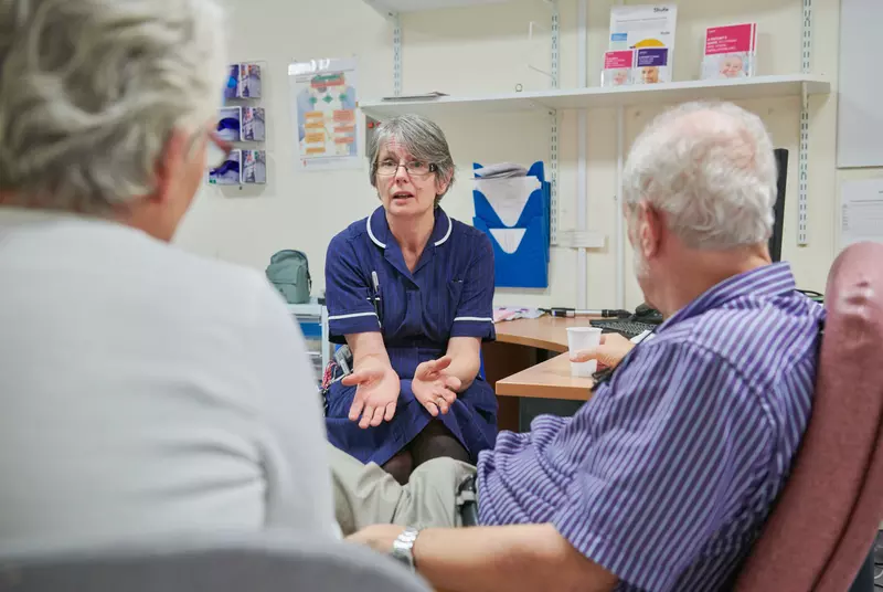 A Parkinson's nurse talking to a person with Parkinson's and his companion in a treatment room.