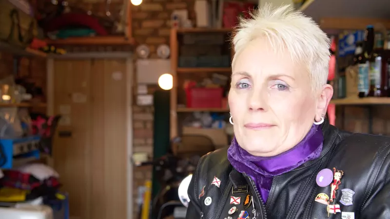 Karen sits in her garage. She has short blonde hair and is smiling to the camera. She wears a purple scarf, and a black leather biker jacket with badges on it.