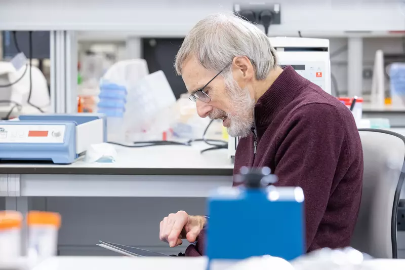 Ken is in a lab. He wears a maroon jumper and is looking down at a tablet computer.