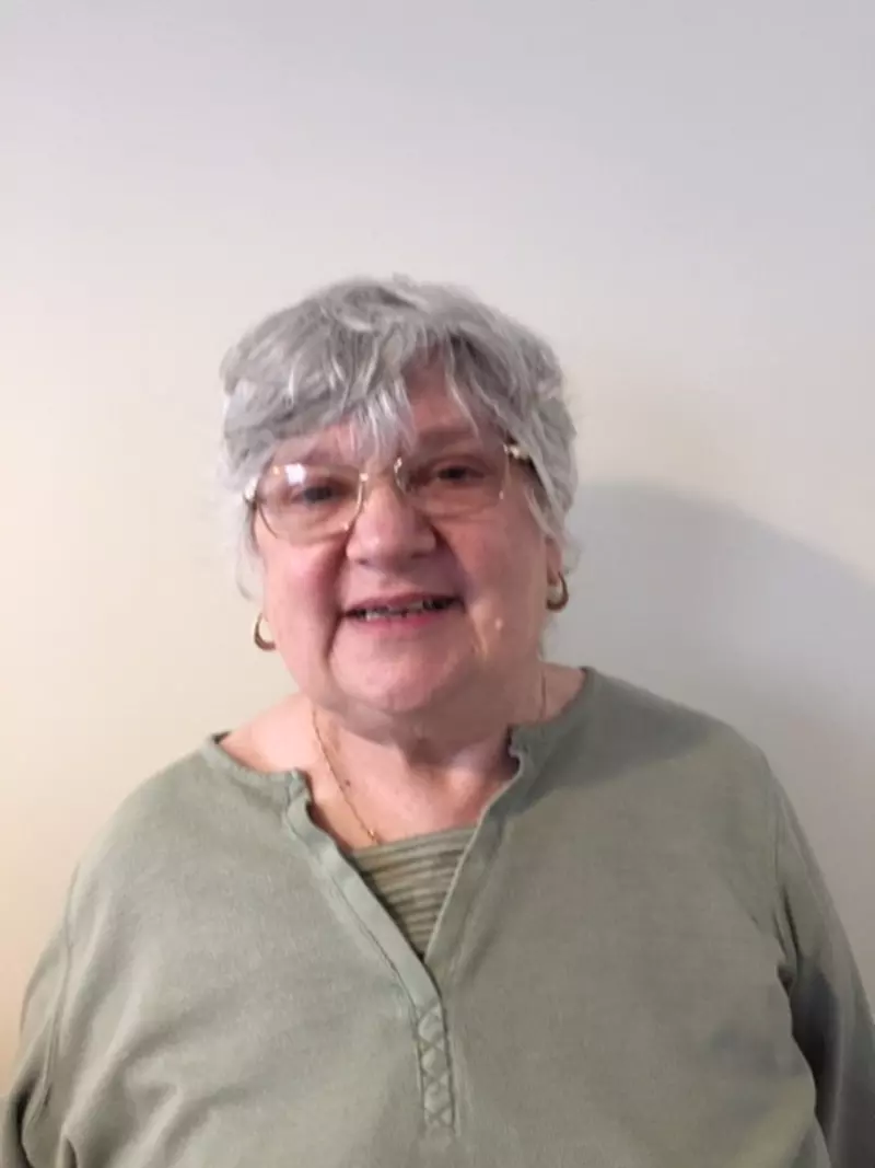 Carole smiling at the camera. She is wearing a khaki green top. She has short grey hair and wears glasses.