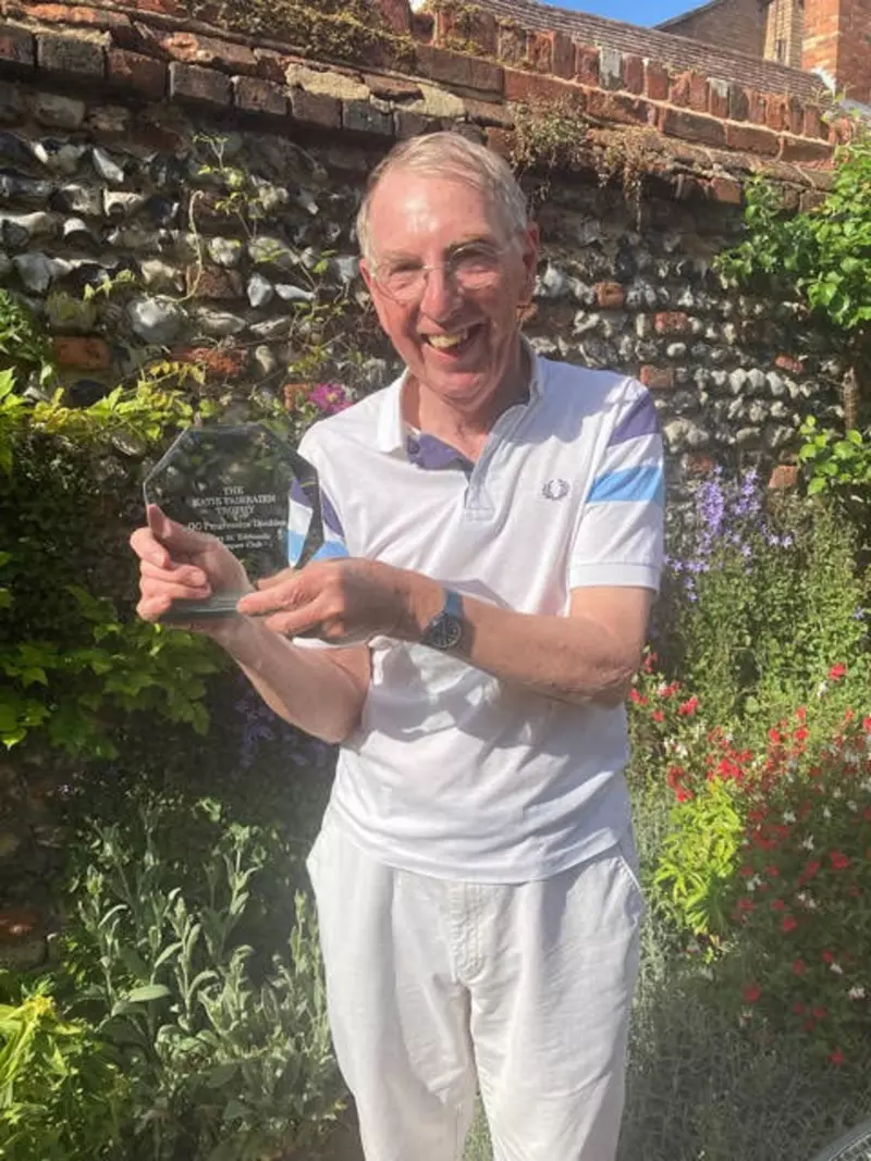 Alan is stood smiling in a walled garden. He is wearing a white polo tshirt and white trousers. He is holding the trophy he won at his croquet club. 