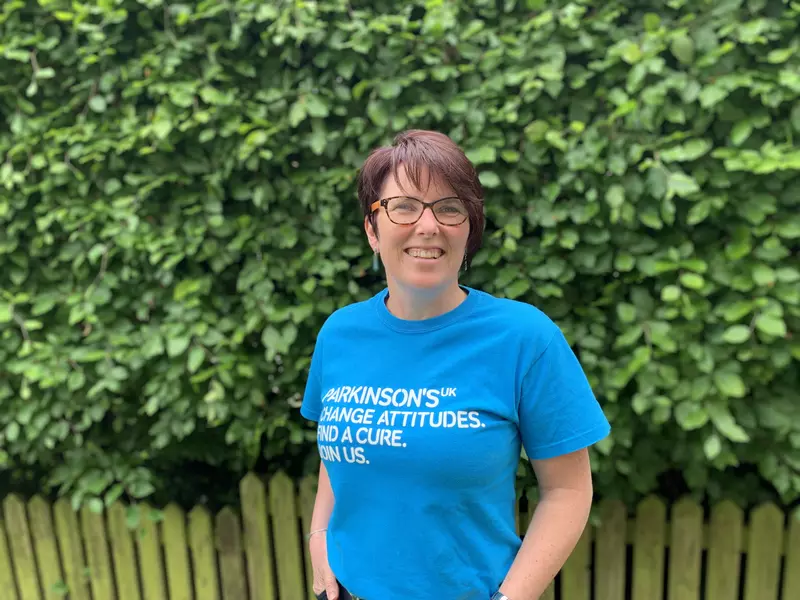 Emma wears her a cyan Parkinson's UK t-short. She has brown hair and wears glasses. She is standing in front of a green hedge.