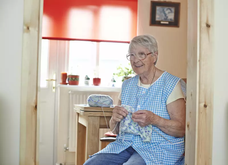 An older lady is knitting at a kitchen table. She has short grey hair and wears glasses. She is wearing jeans, with a yellow tshirt, with a blue tabard.