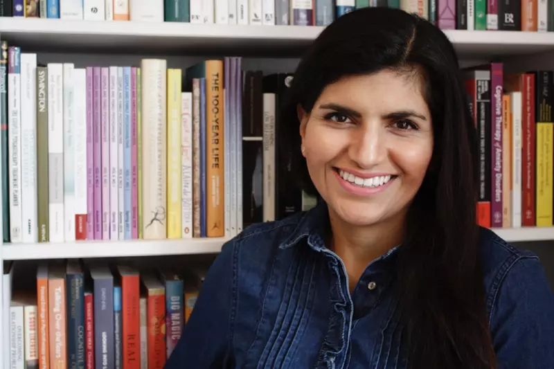 Sabah is smiling to the camera. She has long black hair and is wearing a dark denim shirt. She is in front of a bookcase. 