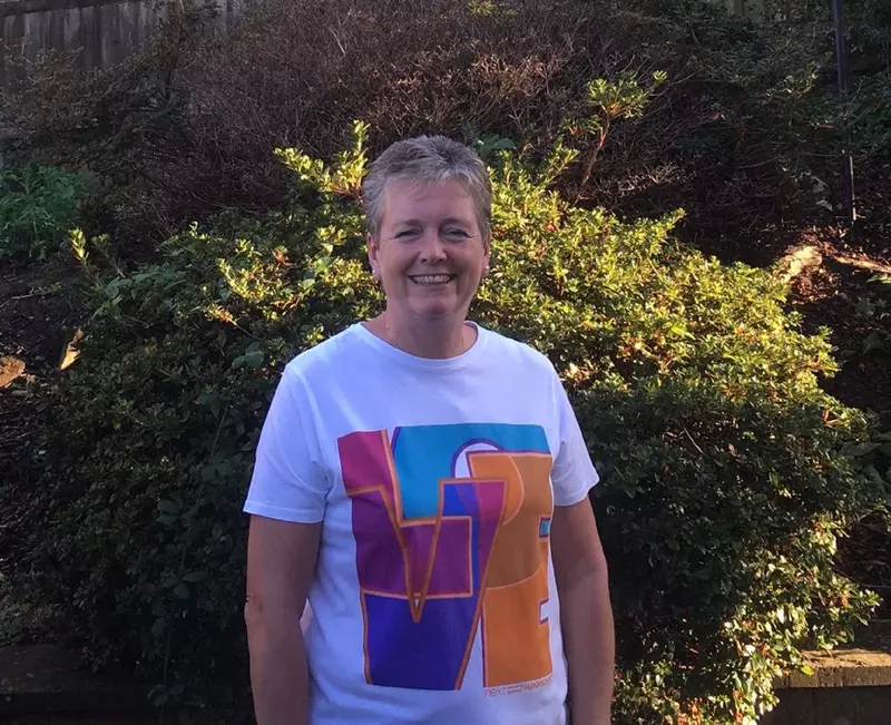 Viv is wearing a white t-shirt wuth a bright design on. She is standing outside in front of a bush. She has short grey hair and is smiling to the camera.