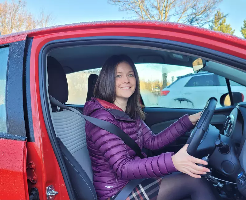 Angela is sat in a red car, holding onto the steering wheel. She is smiling to camera and wearing a purple puffer jacket.