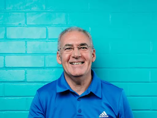 Brian is smiling at the camera. He is wearing glasses and a blue polo tshirt, standing in front of a blue brick wall.
