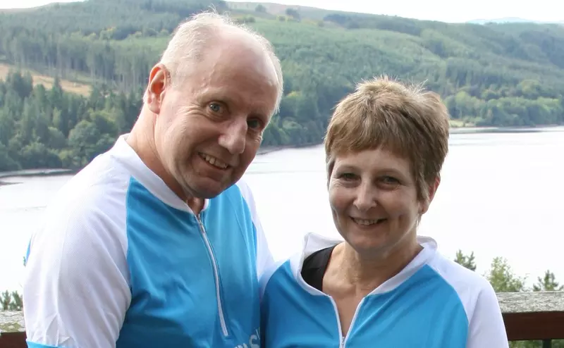 Joan and Fordon smiling to camera, in front of a lake landscape