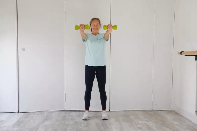 Beth is in sports wear and is holding a dumb bell in each hand, with her arms stretched out in front of her.