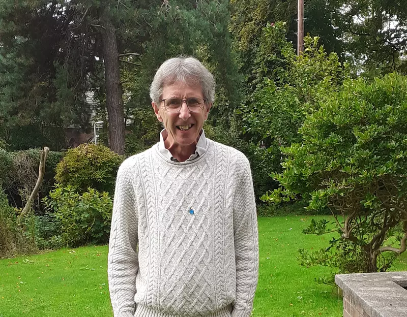 Graham is stood in his garden. He is smiling at the camera and is wearing a cream knitted jumper.