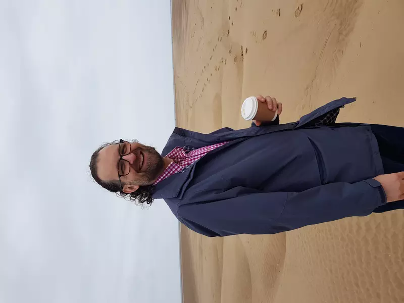 Howard is smiling to camera. He's wearing a blue jacket, holding a coffee cup and is standing on the beach.