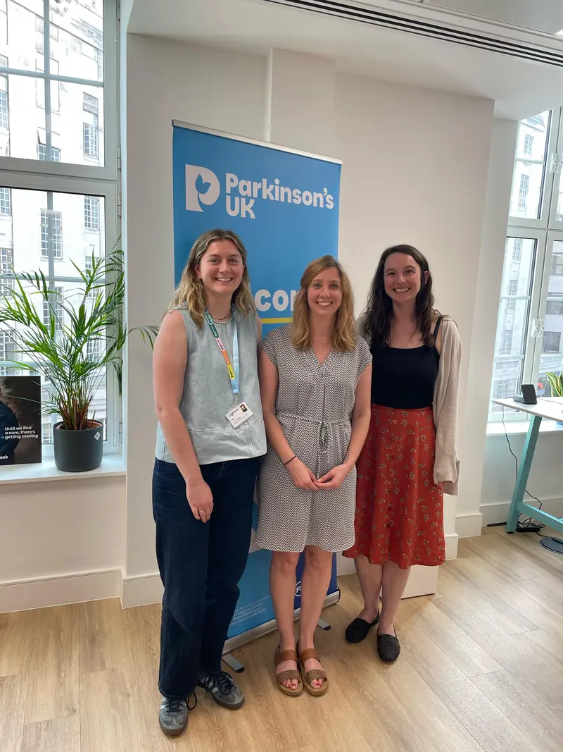 Emma, Katherine and Becky, the Parkinson's UK Research Communications team, standing in front of a Parkinson's UK banner