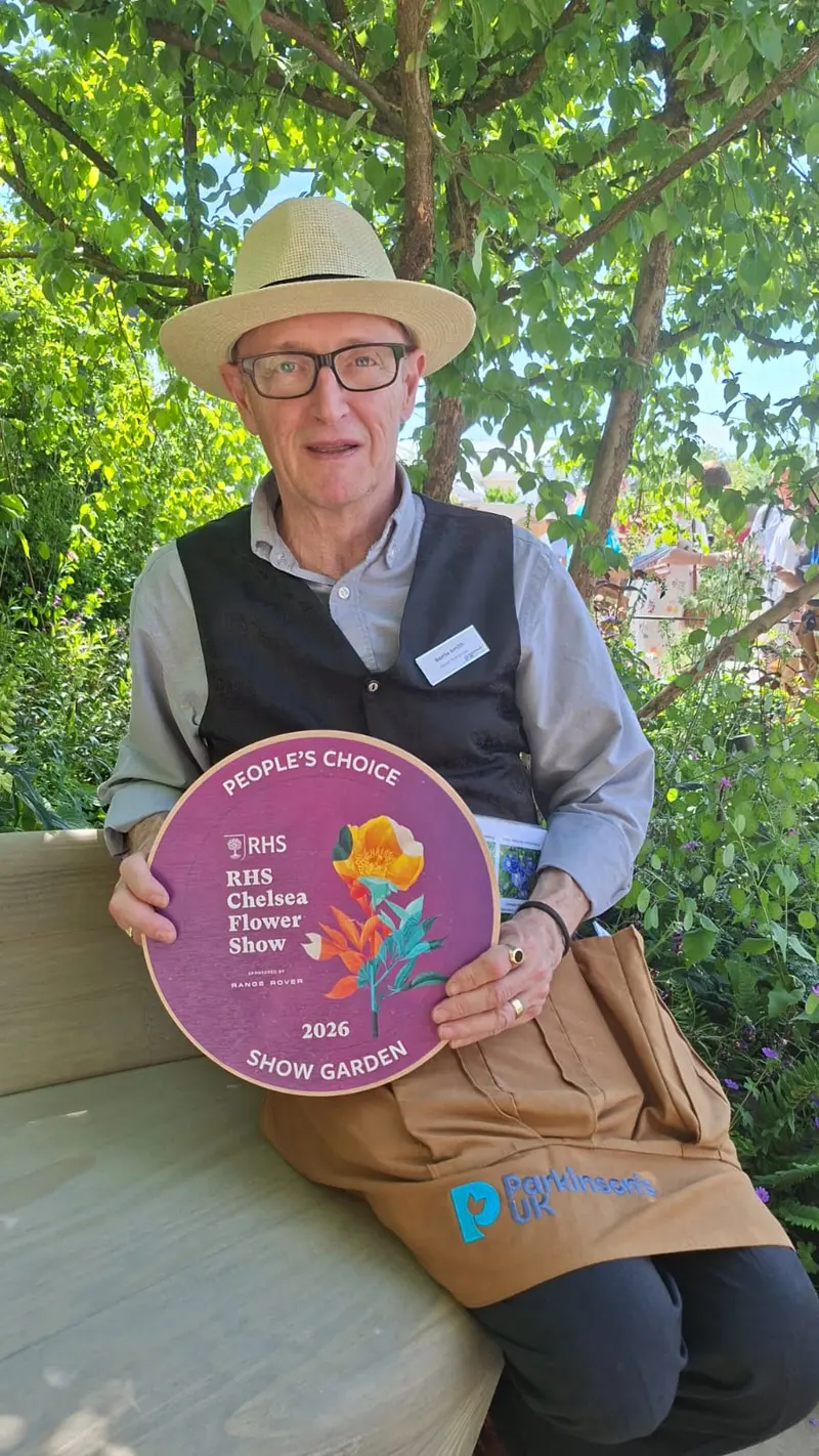 A man in gardening clothes sat in a garden holding an award 