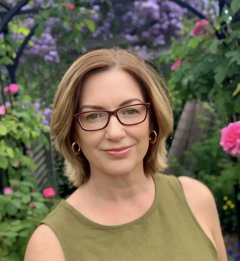 A woman with glasses stands in front of a garden